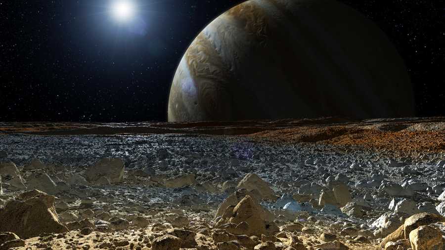 This artist’s concept shows a simulated view from the surface of Jupiter’s moon Europa. Europa’s potentially rough, icy surface, tinged with reddish areas that scientists hope to learn more about, can be seen in the foreground. The giant planet Jupiter looms over the horizon.