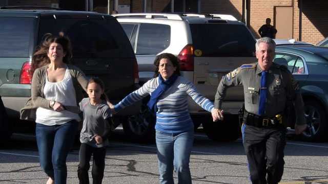 In this photo provided by the Newtown Bee, a police officer leads two women and a child from Sandy Hook Elementary School