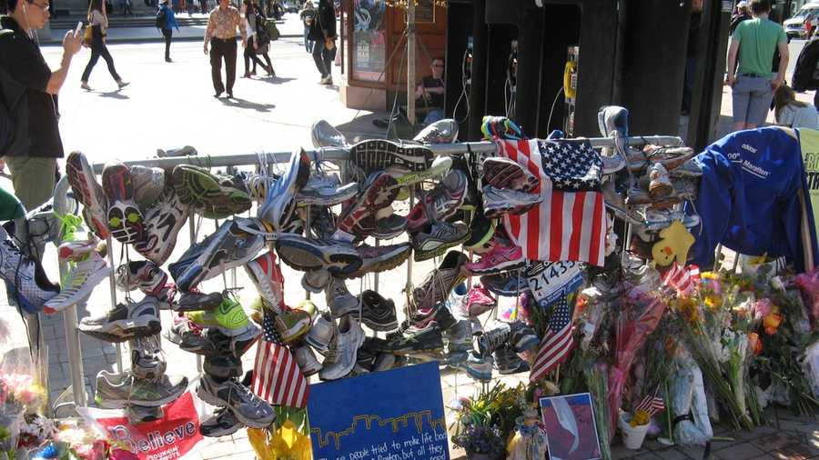A section of fence hung with running shoes, signs, and flowers at the Copley Square memorial. A sign in the foreground reads: "These people tried to make life bad for the city of Boston, but all they can ever do is show just how good those people are. -Stephen Colbert"
