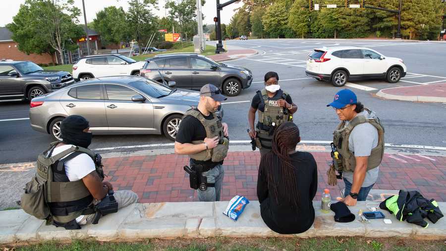 girl sits on ledge with four law enforcement officers standing around her talking in front of an intersection