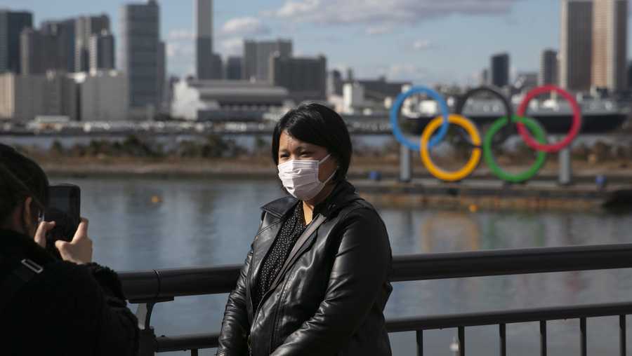 In this Jan. 29, 2020, file photo, a tourist wearing a mask poses for a photo with the Olympic rings in the background, at Tokyo's Odaiba district. Tokyo Olympic organizers repeated their message at the start of two days of meetings with the IOC: this summer's games will not be cancelled or postponed by the coronavirus spreading  neighboring China.