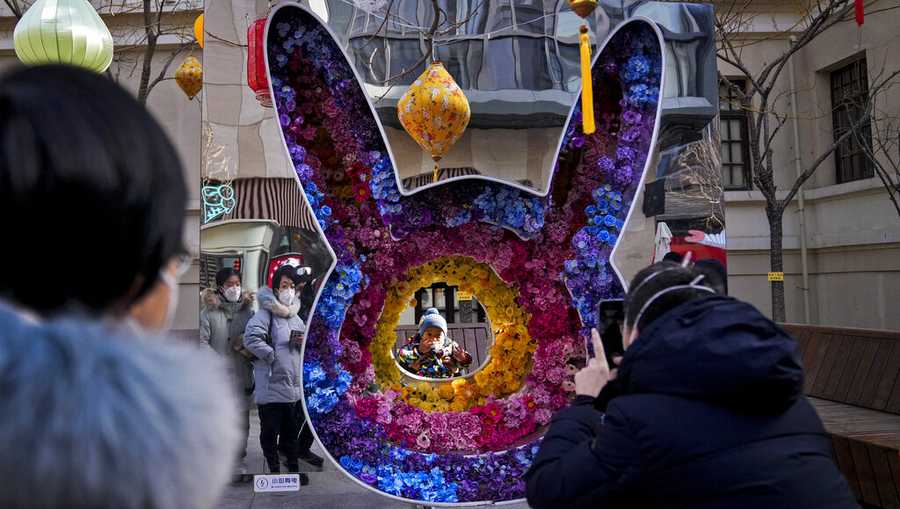 Women wearing face masks watch a toddler pose for a souvenir photo with a rabbit shaped floral decoration at a pedestrian shopping street at Qianmen on the first day of the Lunar New Year holiday in Beijing, Sunday, Jan. 22, 2023. People across China rang in the Lunar New Year on Sunday with large family gatherings and crowds visiting temples after the government lifted its strict "zero-COVID" policy, marking the biggest festive celebration since the pandemic began three years ago. (AP Photo/Andy Wong)