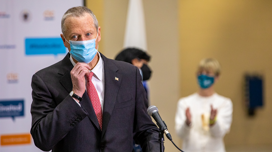 gov. baker touches his mask as he approaches the microphone at a press conference.