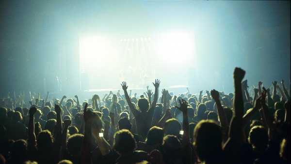 A general view of a rock concert taken from the back for a venue showing the audience in silhouette raising their arms and cheering with bright lights shining from the stage, circa 1980.