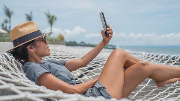 Candid shot of an Indonesian woman taking a selfie during her vacations. She's wearing a straw hat and sunglasses while lying down on a hanging suspended net hammock at seaside restaurant.