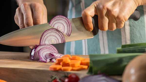 Close-up of female hands cutting onion with knife on chopping board in kitchen.