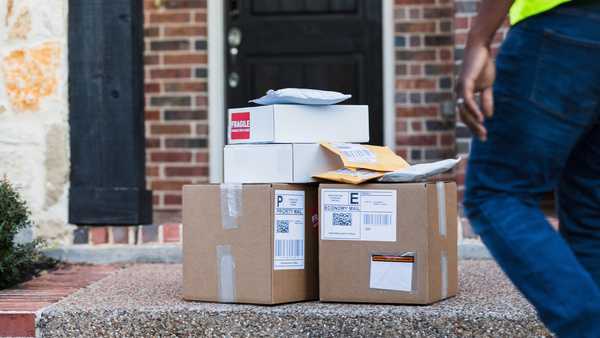 An unrecognizable mail man approaches the stack of boxes and envelopes left on the front porch of the home for pick-up.