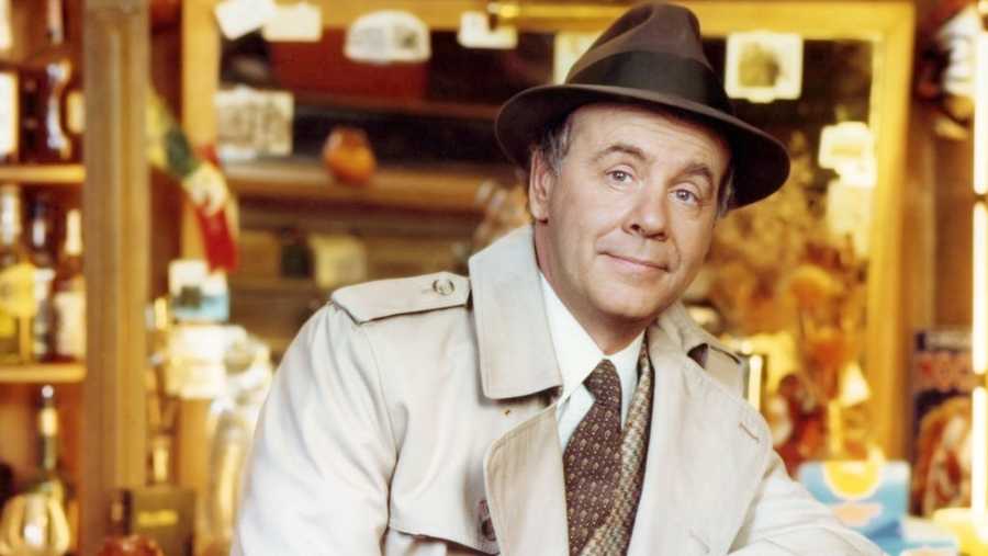 Tim Conway, US actor and comedian, wearing a beige overcoat over a tweed jacket, a white shirt and brown tie, with a brown fedora with a black band, leaning on the back rest of a chair in a studio portrait, circa 1975.