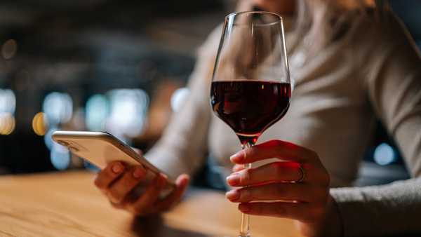 Close-up cropped shot of unrecognizable young woman using smartphone, typing online message sitting at table holding in hand glass of red wine at restaurant. Cute lady chatting with boyfriend.