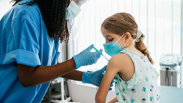 Young girl watching her being injected with COVID-19 vaccine at a medical clinic. African American female nurse or doctor injecting vaccine into caucasian blonde girl patient siiting on examination table in vaccination center.