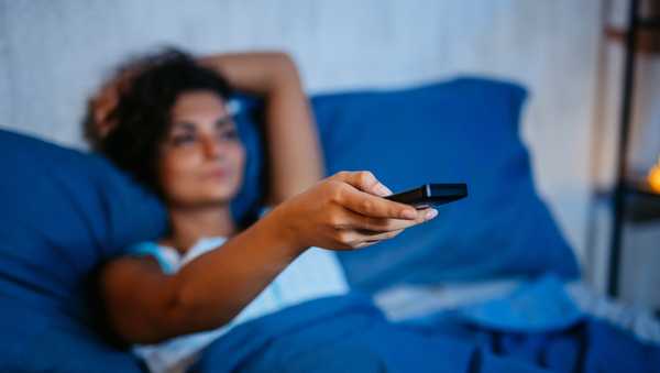 Young woman lying in bed at night, changing channels and watching TV in the bedroom.