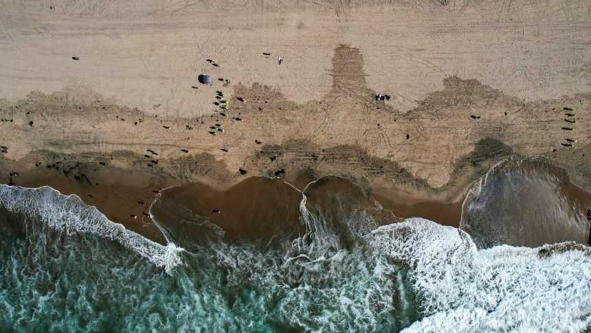 This aerial photo taken with a drone, shows beachgoers as workers in protective suits continue to clean the contaminated beach in Huntington Beach, Calif., on Oct. 11, 2021. Officials were investigating an oil sheen spotted Saturday, Nov. 20, near last month's crude pipeline leak off Southern California's coast. The U.S. Coast Guard said in a statement the oil sheen is about 70 feet by 30 feet and that it has dispatched "pollution responders, aircraft and boats" to investigate.