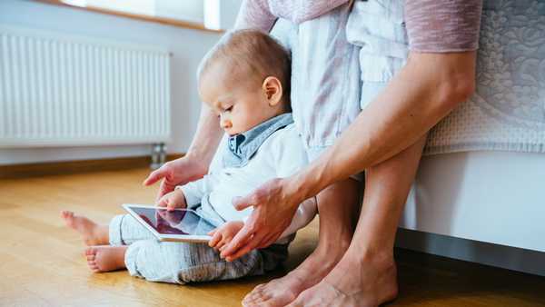 Cute infant baby boy barefoot using a tablet sitting on the flloor, his unrecognizable mother helping him to hold gadget. Real people life concept.