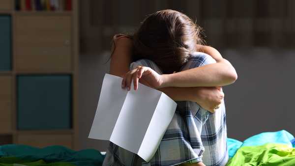 Front view of a single sad teen lamenting sitting on her bed after reading a letter with a dark light in the background