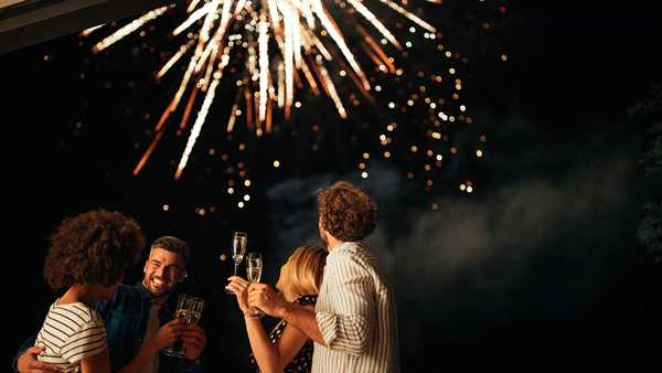 Four friends standing outside, toasting with glasses of wine and watching fireworks