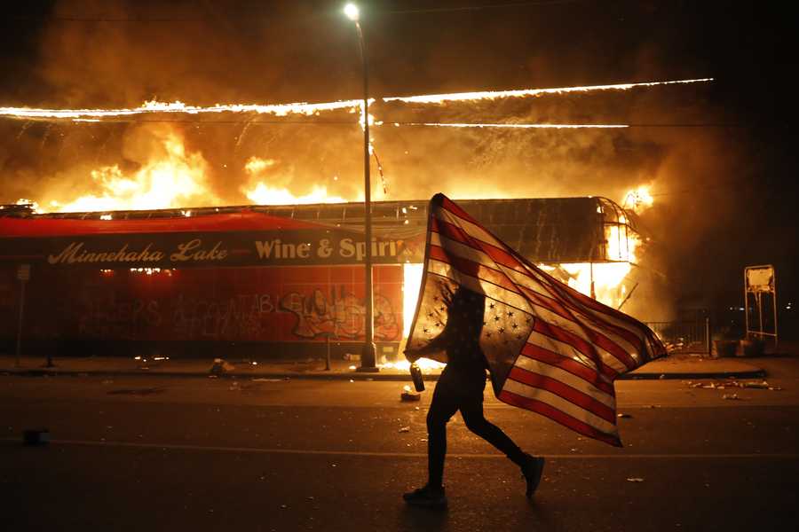A protester carries the carries a U.S. flag upside, a sign of distress, next to a burning building Thursday, May 28, 2020, in Minneapolis.