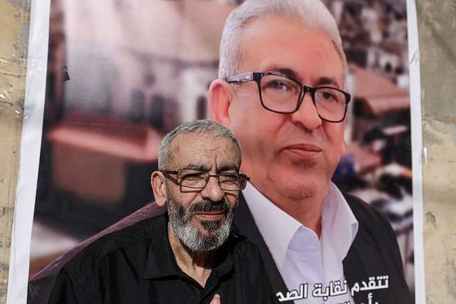 Palestinian journalist Ali al-Samoudi stands in front of a large image of himself taken before he was detained by the Israeli army and held for a year, at his home in the northern city of Jenin, in the Israeli-occupied Palestinian West Bank on May 1, 2026. Sixty-year-old Al-Samoudi was released following one-year in detention in the Israeli Naqab Prison, located in southern Israel. He was released and checked at the Jenin Hospital where he said, "I was 120 kilogrammes; my weight now is barely 60 kilogrammes". Israel has jailed an increasing number of journalists since 2023, the year of Hamas's unprecedented October 7 attack on Israel, which responded with a devastating war in the Gaza Strip. (Photo by JAAFAR ASHTIYEH / AFP via Getty Images)