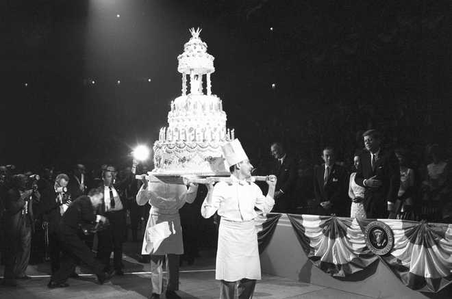 Two chefs carry a multi-tiered birthday cake for President John F. Kennedy, who watches from the side, on May 20, 1962. The celebration took place at a fundraising rally for the Democratic party.