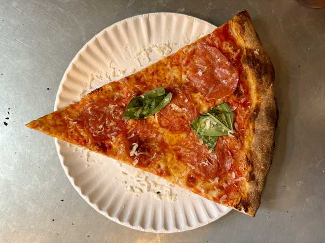 Top-down close-up of a triangular slice of thin-crust pizza with spicy sopressata and basil leaf on the metal counter at a pizzeria in the West Village, New York City