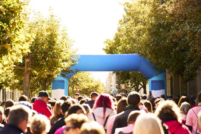 Large crowd of runners and walkers crossing a blue finish arch in sunny urban charity race, celebrating achievement and community energy.