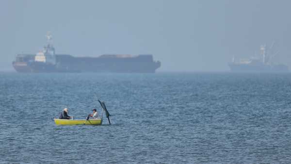 Two men sit in a small boat on the water as cargo ships are anchored in the background in the Strait of Hormuz off Bandar Abbas, Iran, Monday, May 4, 2026.