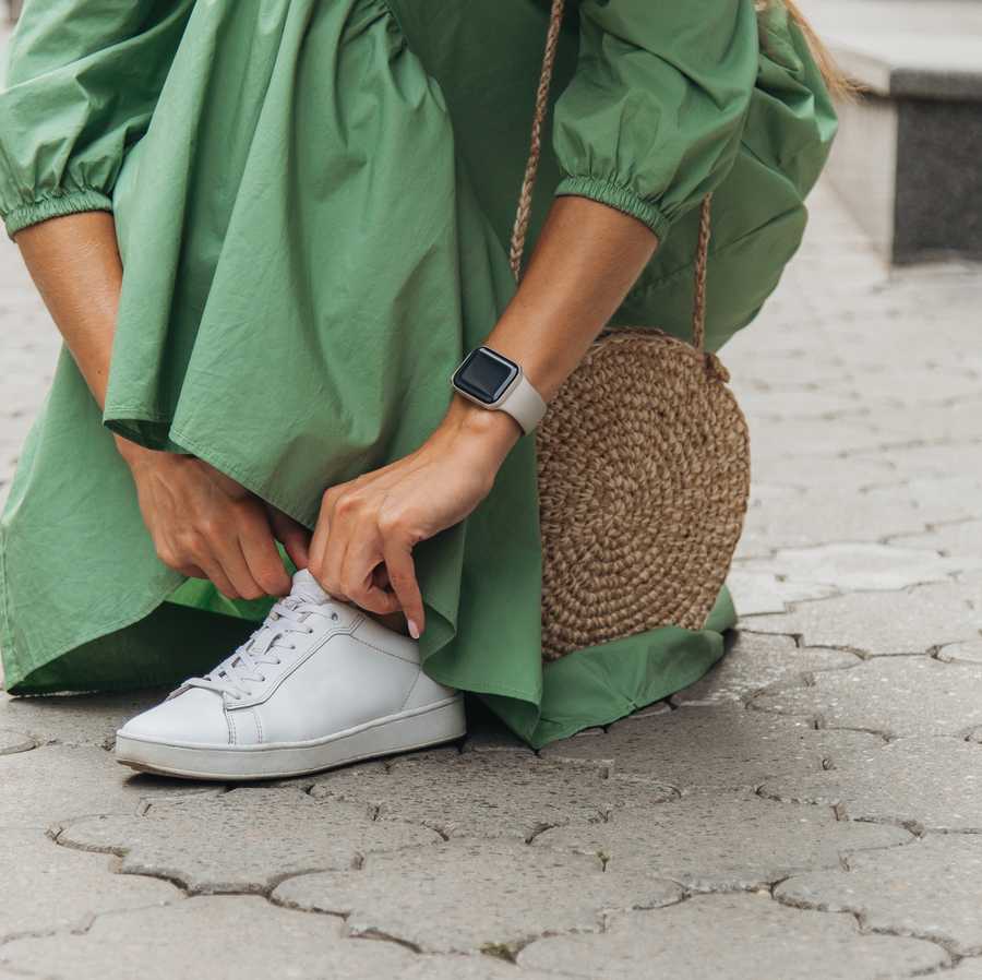 An unrecognizable young woman in a green dress lacing her shoe on a tiled pavement.